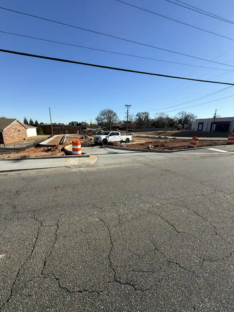 A street corner with newly poured concrete sidewalk, kerb and a detectable warning ramp panel, with orange work-zone barrels and a white pickup truck on the lot behind.