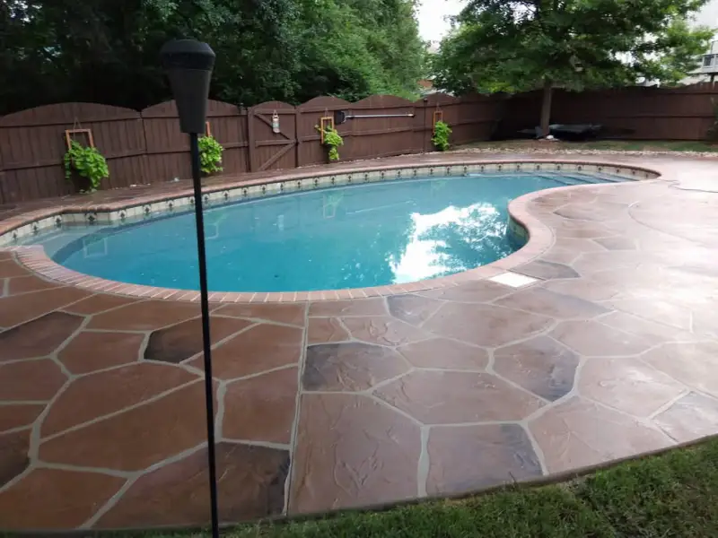 A pool deck surfaced with a hand-troweled flagstone-pattern overlay in warm brown tones, with a brick coping edge around a kidney-shaped pool.