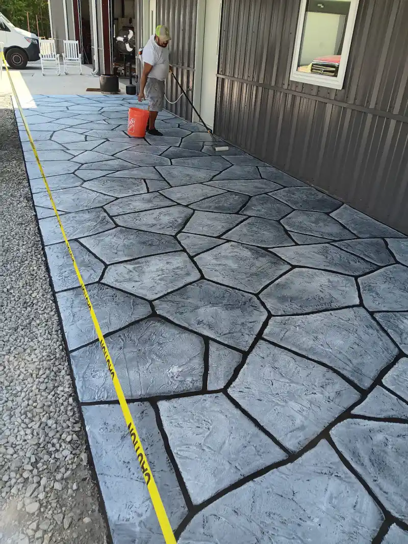 A worker applying a liquid sealer with a long-handled applicator to a dark grey flagstone-pattern overlay walkway beside a metal-clad building.