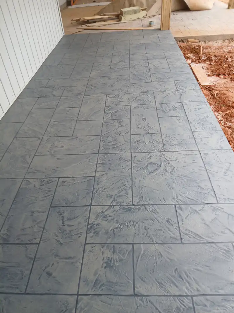 A grey rectangular tile-pattern overlay walkway running beside a white-clad building, with bare red soil and timber offcuts alongside.