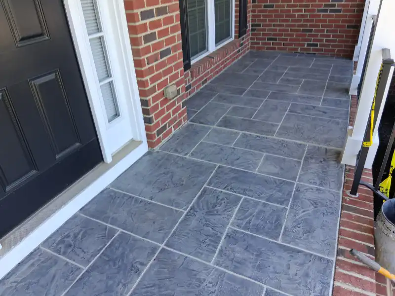 A front porch and entry finished with a dark grey rectangular tile-pattern overlay, next to a black front door and red brick wall.