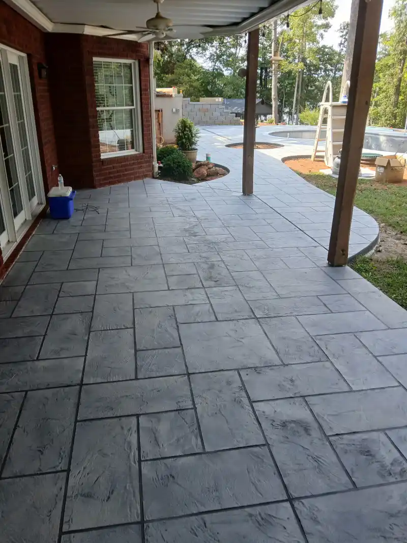 A large patio finished with a tan and grey flagstone-pattern overlay, bordered by a block retaining wall and running to a fire pit at the far end.