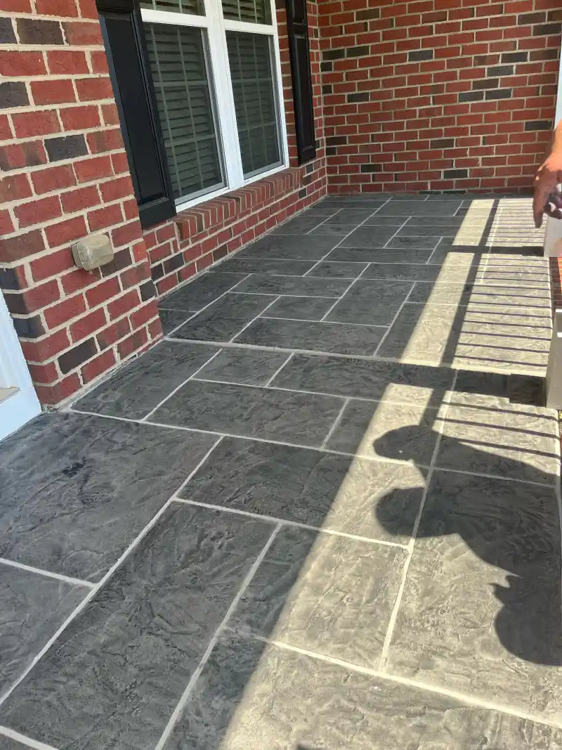 A front porch floor finished with a grey rectangular tile-pattern overlay against a red brick wall.