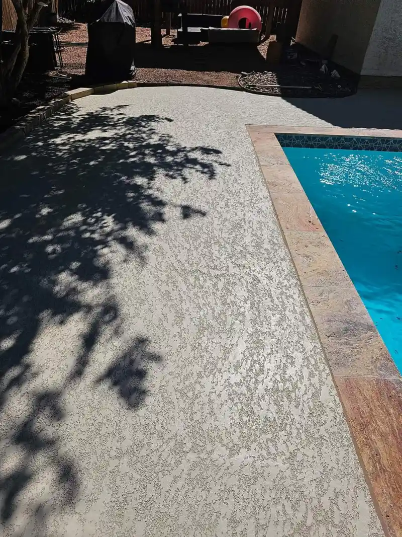 A pool deck finished with a pale knockdown-texture coating, meeting stone coping at the edge of a swimming pool.