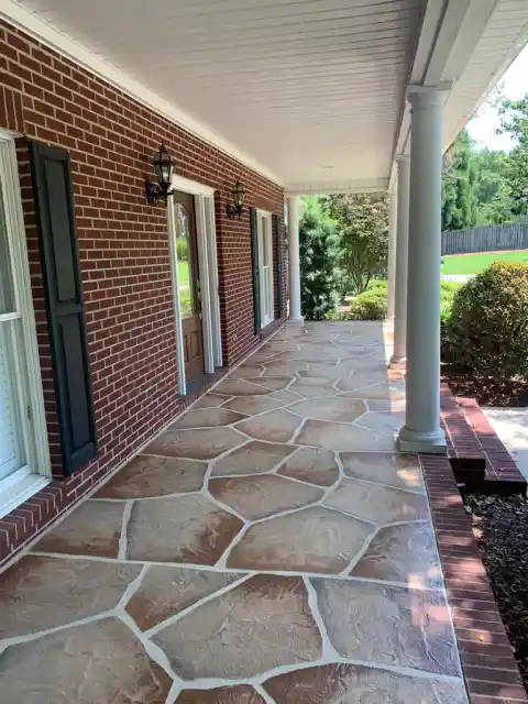 A covered porch at a red brick house finished with a tan and grey flagstone-pattern overlay, framed by white columns.