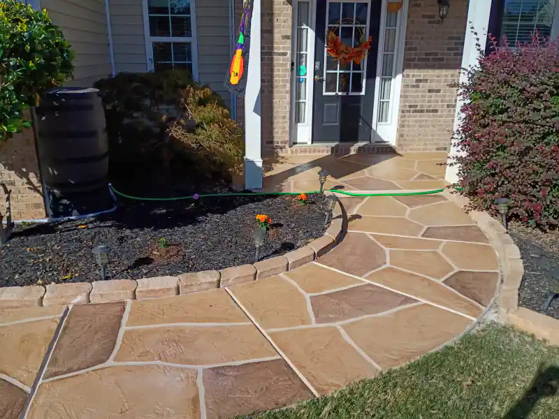 A front walkway finished with a tan flagstone-pattern overlay, curving past a planting bed with a stone border to a house entrance.