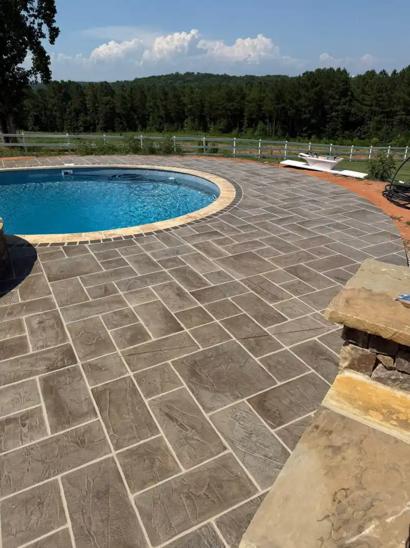 A curved pool deck finished with a brown and grey rectangular tile-pattern overlay, with a white fence and wooded countryside beyond.