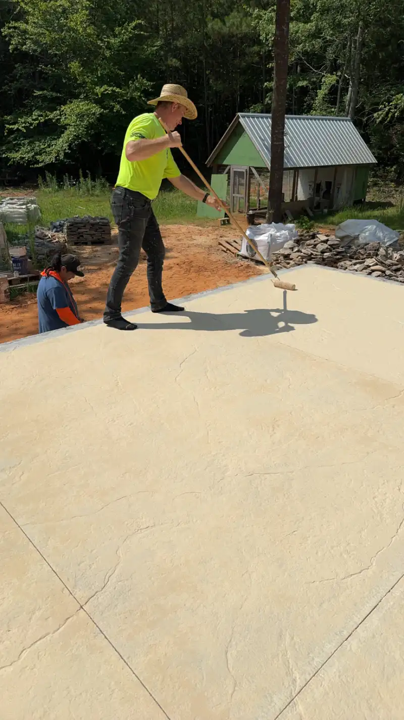 A worker applying a coating with a long-handled roller across a large tan flagstone-textured slab, with a second worker kneeling at the slab edge.