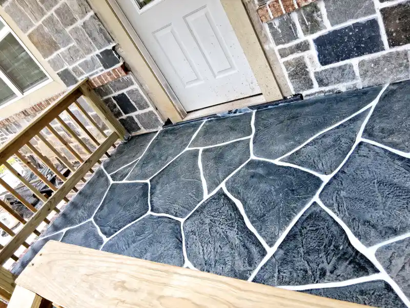 A porch floor finished with a dark charcoal flagstone-pattern overlay with white jointing, at the door of a stone-clad house with a timber handrail.