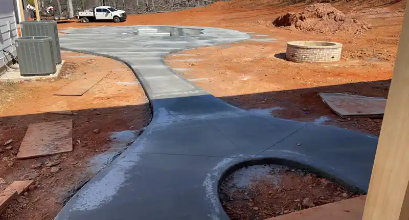 A curved concrete walkway joining a driveway to a patio area, with a brick fire pit ring and two air conditioning units alongside.