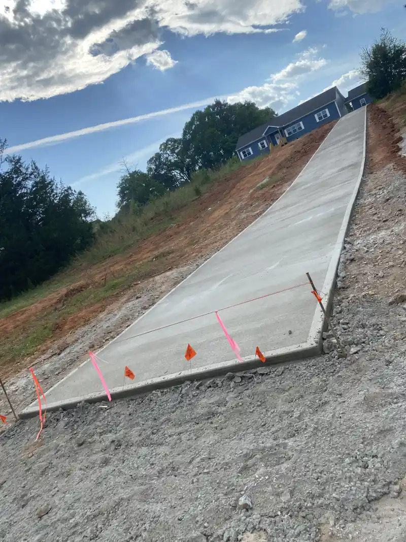 A long concrete driveway sloping uphill toward a two-storey house, with survey marker flags along the edge.