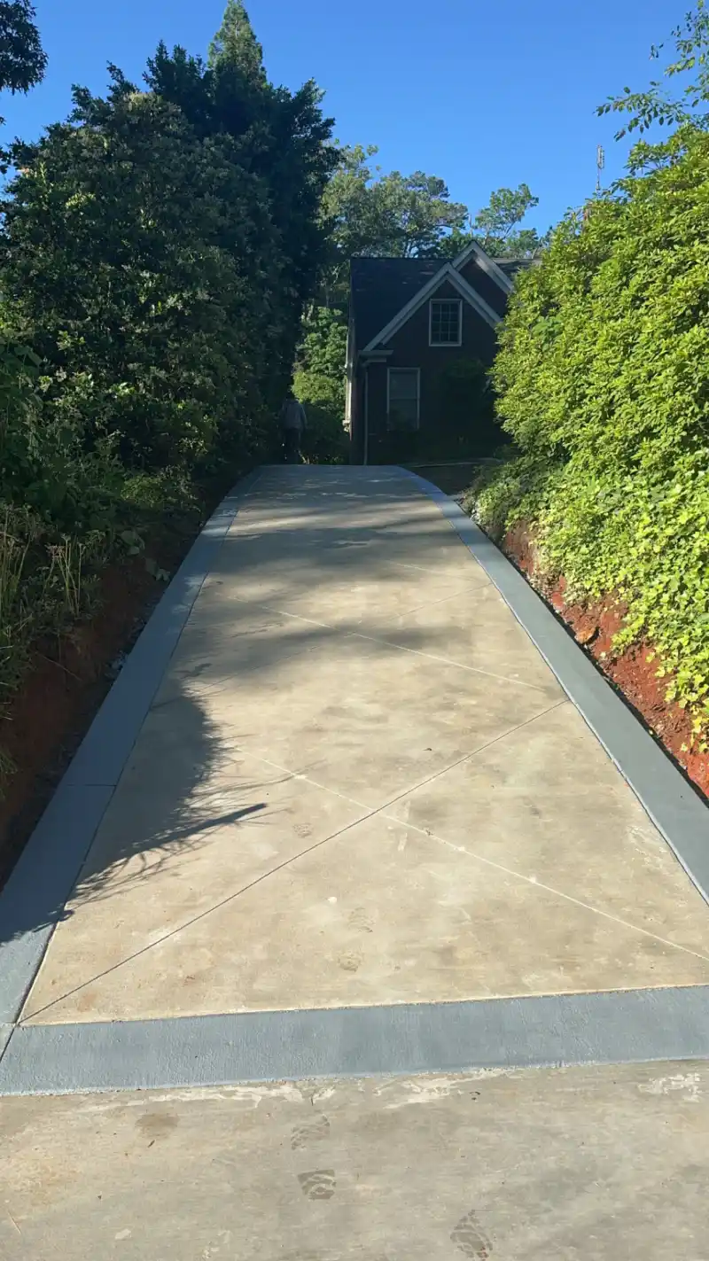 A concrete driveway with a contrasting bordered edge running between a clipped hedge and a planted bank toward a brick house.