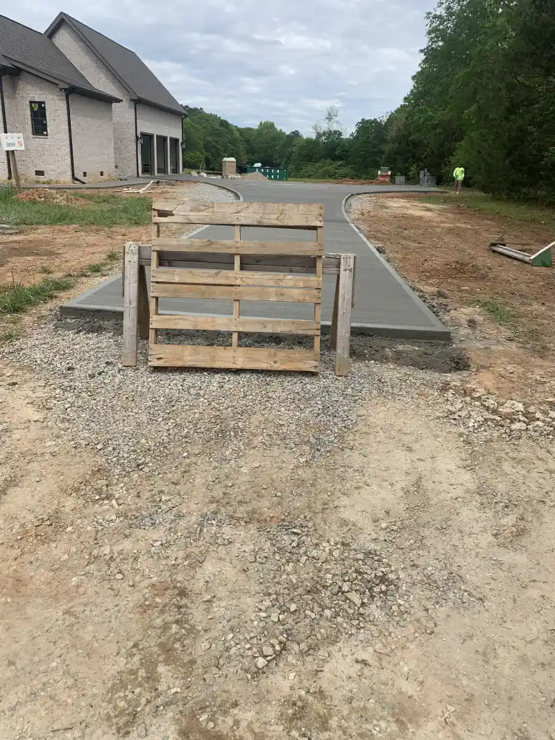 A newly poured concrete driveway at a large brick house, with a timber pallet propped across the entrance as a barrier.