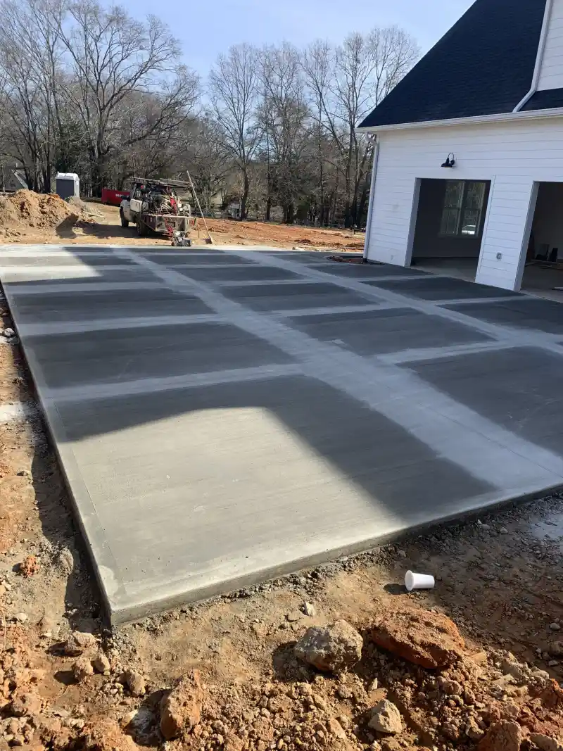A large freshly finished concrete parking pad beside a white house with open garage bays.