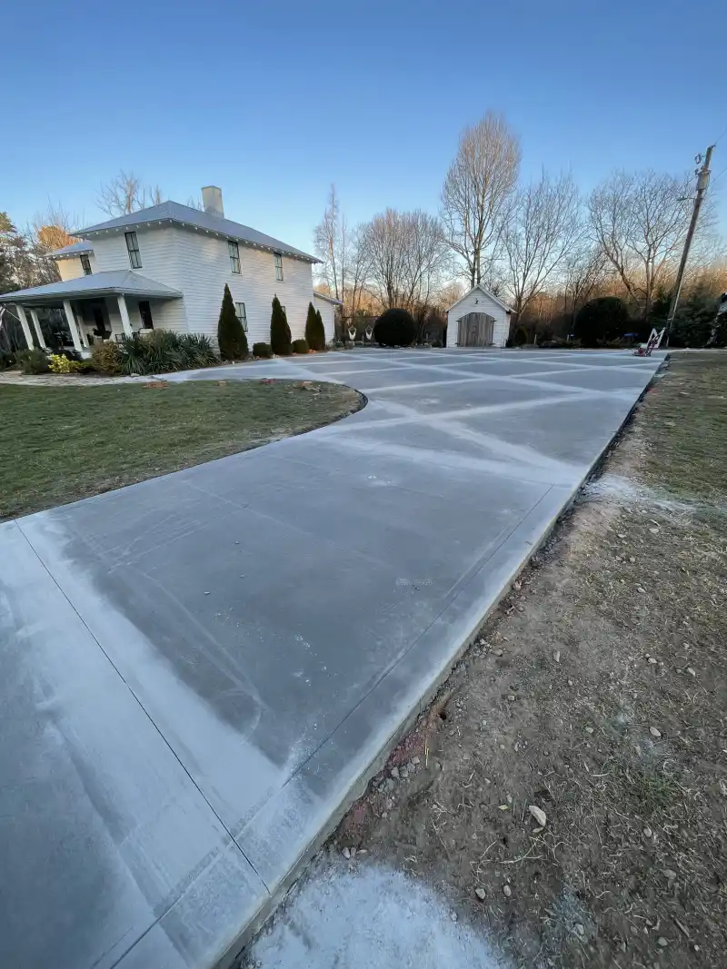 A wide concrete driveway leading to a farmhouse, photographed in low evening sunlight.