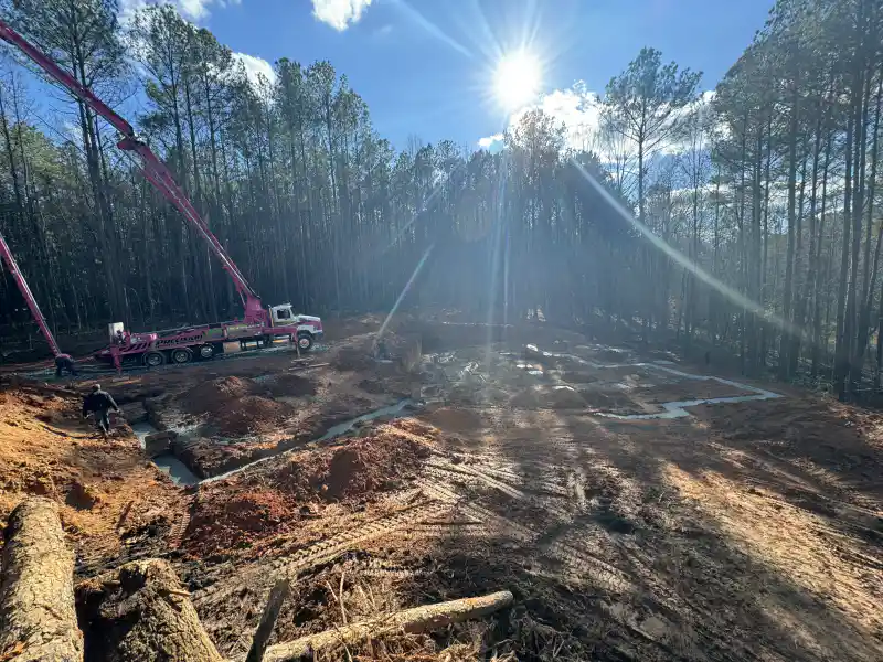 Concrete footings being poured across a cleared wooded lot, with a boom pump truck reaching over the excavation and strong sun flare in the frame.