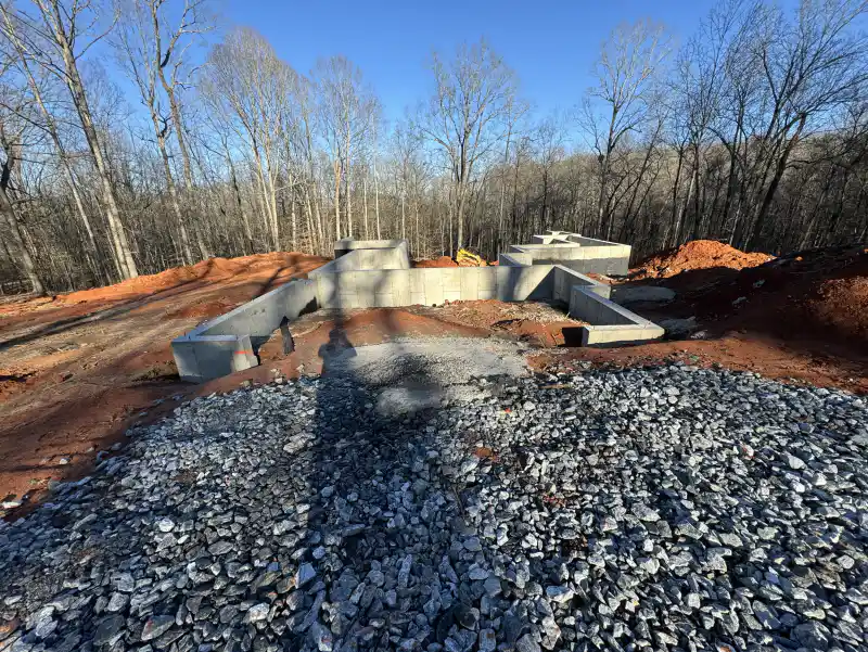 Stepped concrete foundation walls on a cleared lot, with crushed stone in the foreground and bare winter woodland behind.