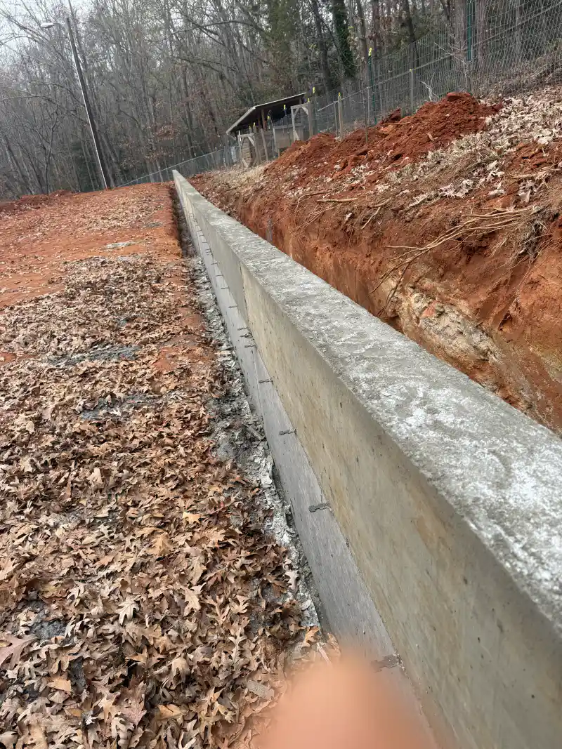 A poured concrete retaining wall running along an excavated clay bank beside a chain-link fence.