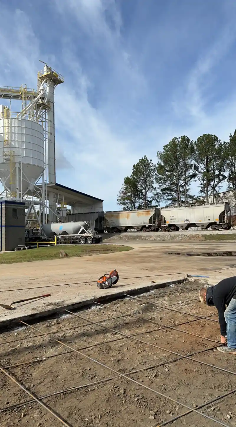 A worker tying steel reinforcing bar at an industrial site with storage silos and rail infrastructure behind.
